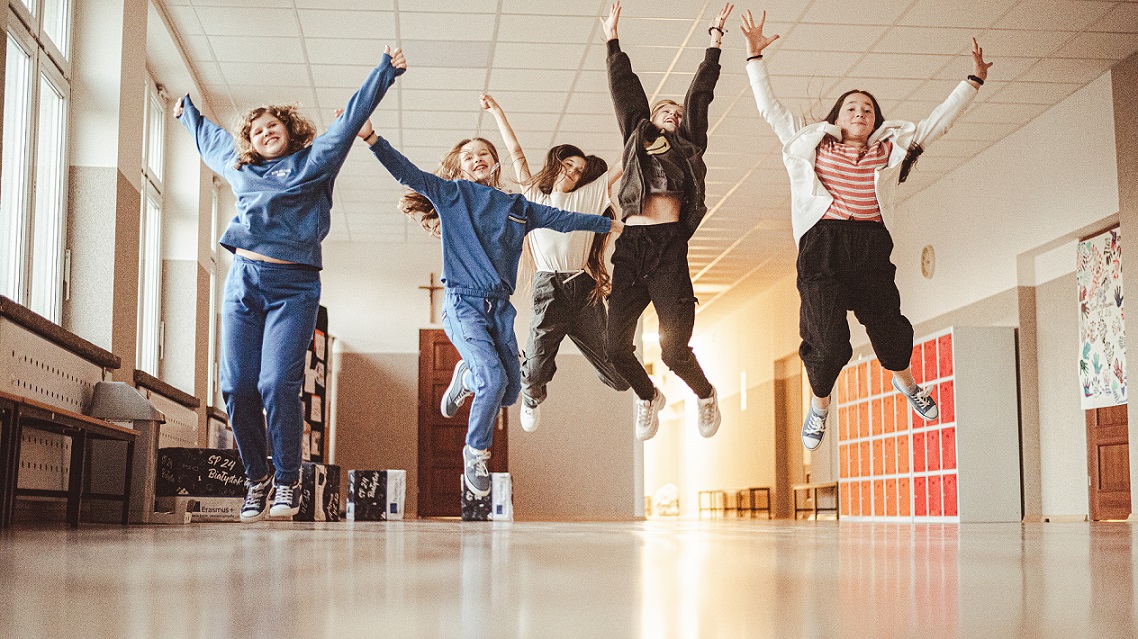 Pięć dziewczynek skacze do góry na korytarzu szkolnym.