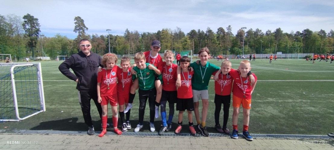 Grupa chłopców w czerwonych strojach sportowych pozuje razem z trenerem na boisku do piłki nożnej..jpg