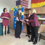 Uczennica odbiera dyplom od kobiety ubranej w bordową marynarkę. W tle flagi Niemiec i USA.jpg