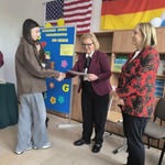 Uczennica odbiera dyplom od kobiety ubranej w kolorowa marynarkę. W tle flagi Niemiec i USA.jpg