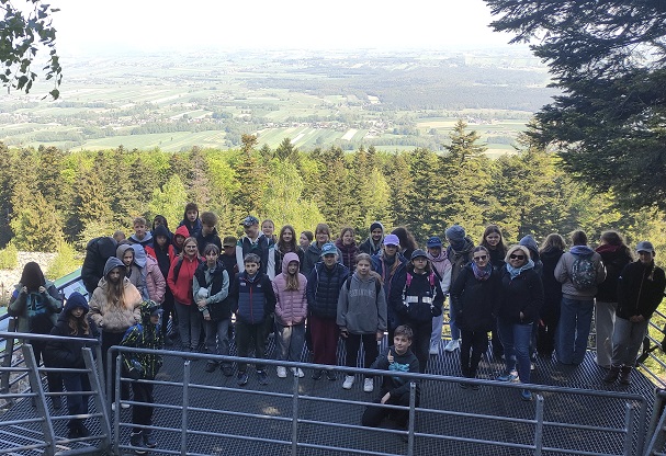 Grupa młodzieży i osób dorosłych stoi na kładce na tle panoramy na zielone pola i lasy.jpg