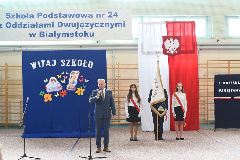 Dyrektor szkoły przemawia na tle napisów na niebieskim tle. Za nim stoi poczet sztandarowy na tle flagi biało-czerwonej..jpg