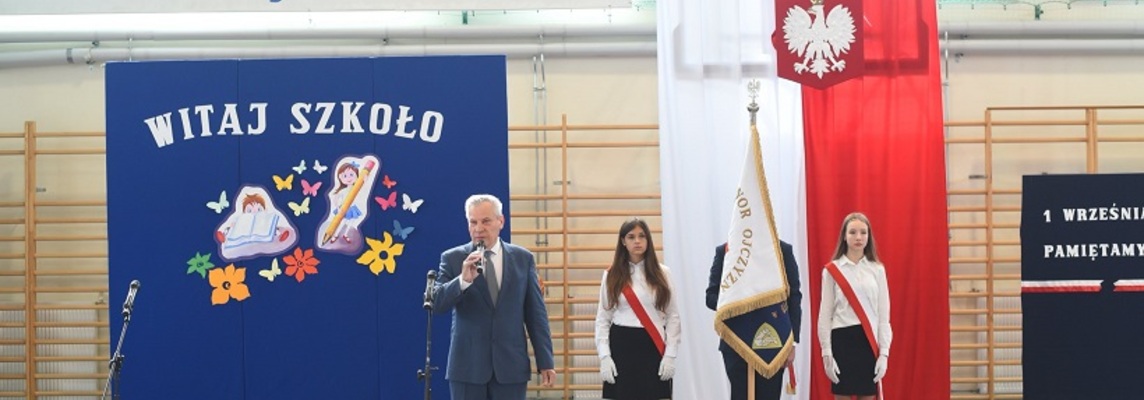 Dyrektor szkoły przemawia na tle napisów na niebieskim tle. Za nim stoi poczet sztandarowy na tle flagi biało-czerwonej..jpg