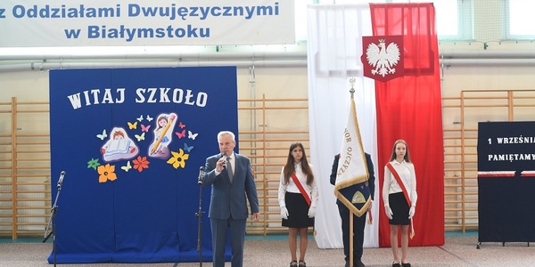Dyrektor szkoły przemawia na tle napisów na niebieskim tle. Za nim stoi poczet sztandarowy na tle flagi biało-czerwonej..jpg