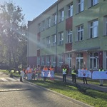 Grupa dzieci w kamizelkach odblaskowych stoi przed budynkiem szkoły i trzyma plakaty.jpg