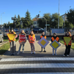 Na chodniku przed ogrodzeniem stoi sześcioro dzieci w kamizelkach odblaskowych trzymających kartonowe znaki drogowe..png