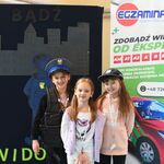Trzy dziewczynki_ dwie w czapkach policyjnych pozują do zdjęcia na tle granatowego plakatu z szarym odwróconym chłopcem..jpg