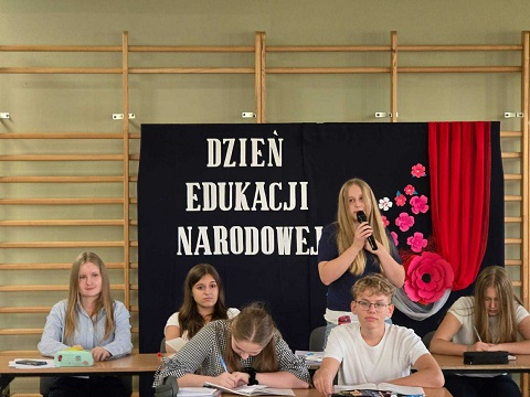 Grupa uczniów siedząca w ławkach_  jedna z nich mówi do mikrofonu. W tle granatowa tablica z kwiatami..jpg