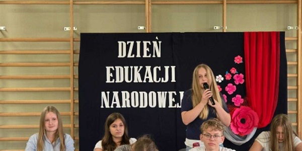 Grupa uczniów siedząca w ławkach_  jedna z nich mówi do mikrofonu. W tle granatowa tablica z kwiatami..jpg