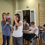 Grupa uczniów siedząca w ławkach_  jedna uczennica mówi do mikrofonu_ druga trzyma mazak w dłoni.jpg
