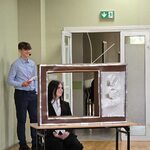 Uczennica ubrana w galowy strój siedzi za atrapą telewizora. Z tyłu stoi chłopak w niebieskiej koszuli..jpg
