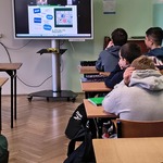 Widok monitora z wyświetlanym tekstem_ przed nim siedzący tułem uczniowie w ławkach.jpg