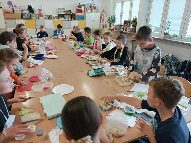 Grupa dzieci siedzi przy dużym stole w klasie i przygotowuje kanapki z produktów spożywczych..jpg