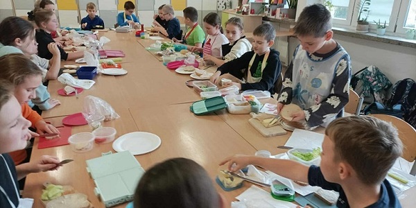 Grupa dzieci siedzi przy dużym stole w klasie i przygotowuje kanapki z produktów spożywczych..jpg
