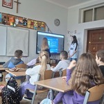 Grupa uczniów siedzi w ławkach w klasie i patrzy na dwie dziewczyny stojące przy monitorze.jpg