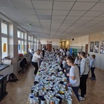 Korytarz szkolny wypełniony dużą ilościa kubków ustawionych na stołach oraz dziećmi stojącymi przy nich.jpg