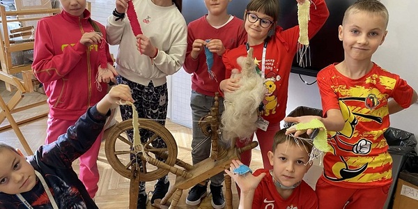 Grupa dzieci ubrana w czerwone bluzy stoi przy kołowrotku z utkanymi paskami w rękach.jpg