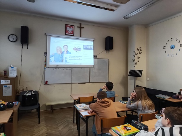 Grupa uczniów siedzi w ławkach klasowych i ogląda prezentację na ekranie.jpg