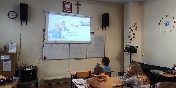 Grupa uczniów siedzi w ławkach klasowych i ogląda prezentację na ekranie.jpg