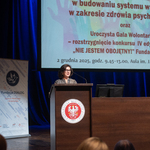 Kobieta przemawia na mównicy z logo orła, w tle ekran z napisami