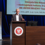Mężczyzna przemawia na mównicy z logo orła, w tle ekran z napisami
