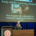 Kobieta przemawia na mównicy z logo orła, w tle ekran z napisami i zdjęciem rąk.