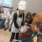 Dziewczynka w czapce piekarza na głowie rozdaje siedzącym w kręgu dzieciom ciastka.jpg