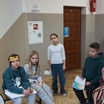 W centrum stoi dziewczynka w białej bluzce i spodniach_ obok niej na ławkach siedzą trzej chłopcy.jpg