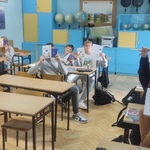 Grupa uczniów siedzi w ławkach w klasie i trzyma w górze kartki.jpg