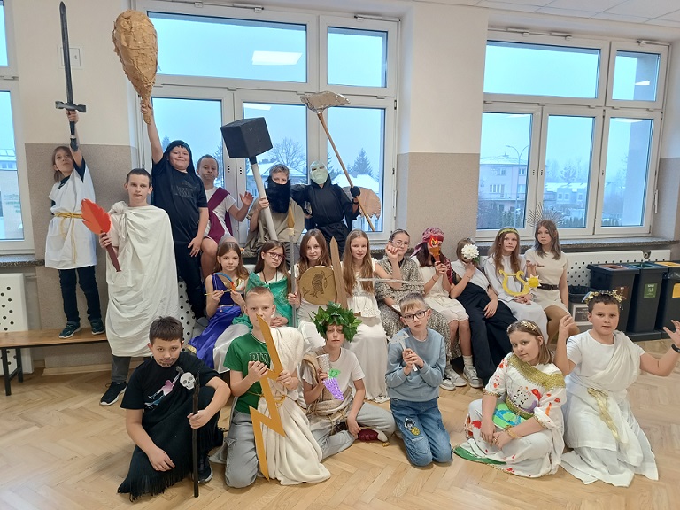 Grupa uczniów stoi lub siedzi na kanapie i podłodze na tle okna i prezentuje atrybuty postaci mitologicznych .jpg