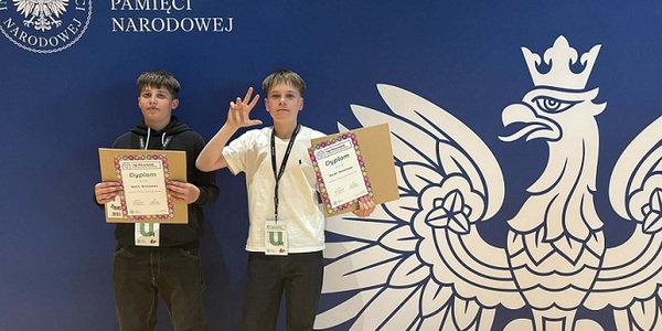 Dwóch chłopców stoi z dyplomami na tle baneru IPN z białym orłem.jpg