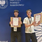 Dwóch chłopców stoi z dyplomami na tle baneru IPN z białym orłem..jpg