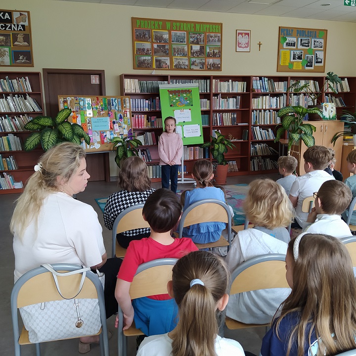 Uczennica stoi w bibliotece przed uczniami siedzącymi na krzesłach.jpg