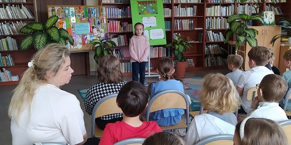 Uczennica stoi w bibliotece przed uczniami siedzącymi na krzesłach.jpg