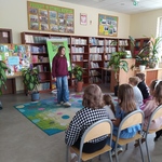 Uczennica stoi w bibliotece na kolorowym dywanie przed uczniami siedzącymi na krzesłach.jpg