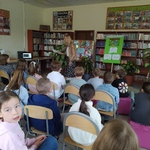 Dzieci siedzą na krzesłach w bibliotece i patrza w kierunku kobiety trzymjącej książkę.jpg