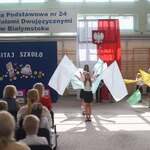 Grupa dzieci tańczy z kolorowymi flagami na tle napisu i flagi polskiej z godłem.jpg
