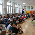 Grupa uczniów siedzi na podłodze holu szkolnego i słucha nauczycieli_ którzy czytają.jpg