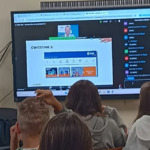 Uczniowie w ławkach obserwują monitor i prezentowane plansze z zadaniami.jpg