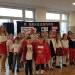 Uczniowie klasy ubrani w storoje biało-czerwone wraz z nauczycielką stoja przy tablicy z wystawą.