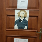 Brązowe drzwi klasy ozdobione plakatem z podobizną osoby oraz napisem.