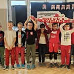 Uczniowie klasy ubrani w stroje biało-czerwone stoją przy tablicy z wystawą. W górze trzymają biało-czerwony szalik.