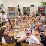  Uczniowie siedzą przy dużym stole i jedzą pizzę.