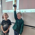Dwójka uczniów stoi przy tablicy interaktywnej, jeden z nich podnosi figurkę do góry.