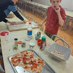Dwóch chłopców stoi przy stole, jeden wałkuje pizzę a drugi wkłada do ust pomidora.