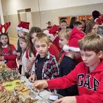 Przed stołami ze smakołykami i dekoracjami świątecznymi stoją ubrane w mikołajkowe stroje dzieci.