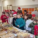 Na szkolnym korytarzu stoją stoły zasatwione smakołykami oraz dekoracjami świątecznymi. Wokół stoją ubrane w mikołajkowe stroje dzieci. 