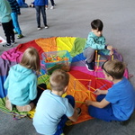 Grupa dzieci siedzi na kolorowej chuście i układa kolorowe patyczki 