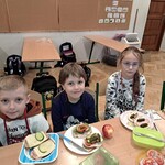 Trójka dzieci siedzi przy stolikach na których stoją talerze z kanapkami.