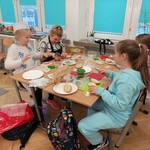 Dzieci siedza przy stole w klasie, leżą na nim talerze z kanapkami. Dzieci robią kanapki.